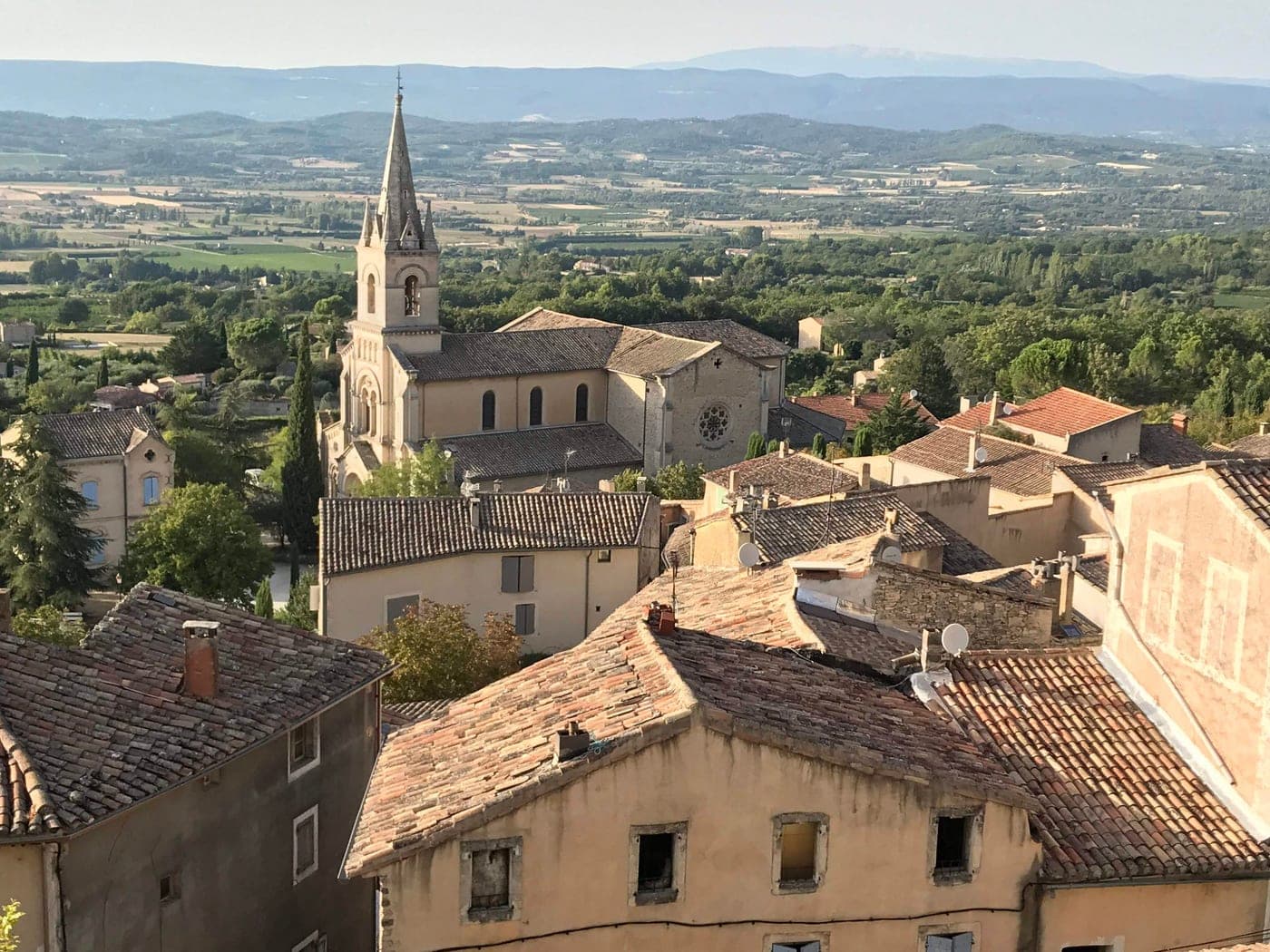 Village de pierre aux toits de tuiles et clocher, dominant une vallée boisée